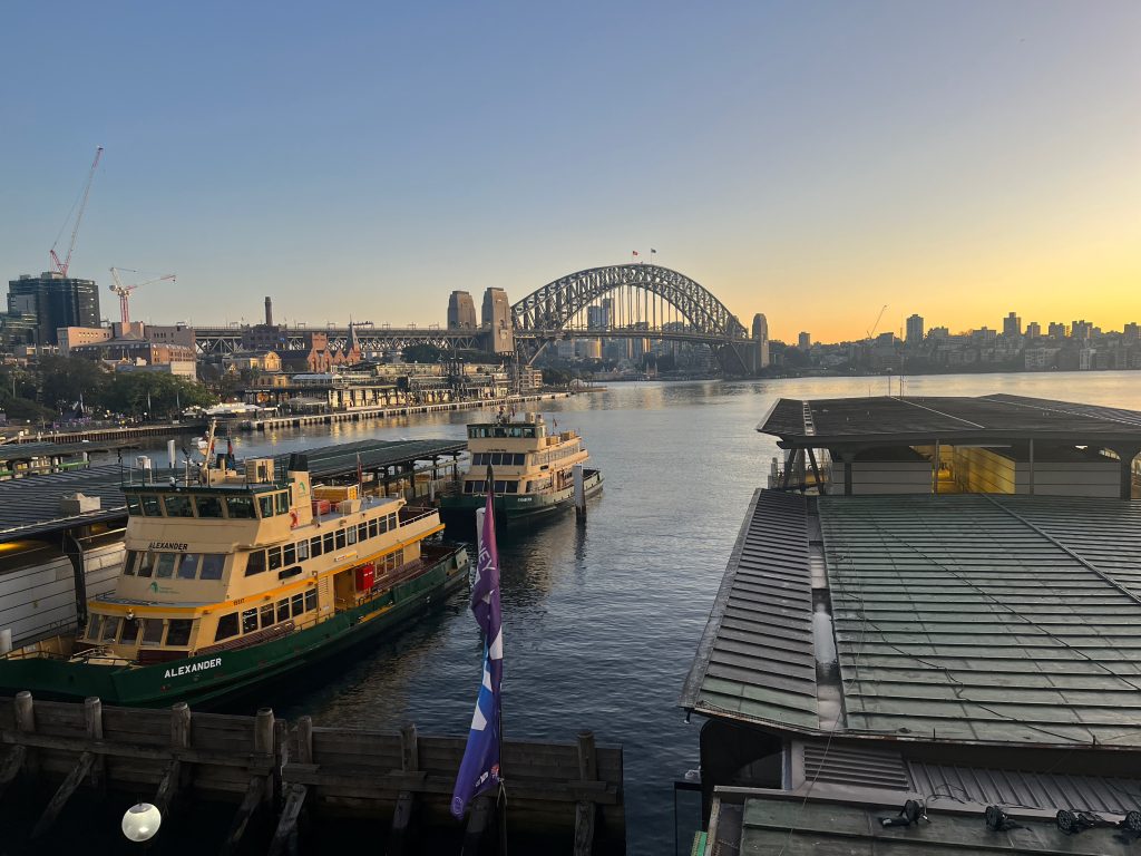A view from the train station at Circular Quay overlooking Sydney Harbour at sunrise, a ferry is in the foreground and behind it, the Sydney Harbour Bridge